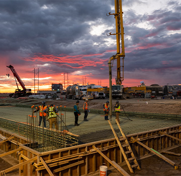 Construction workers on a rebar grid at dusk with a concrete pump and crane under a dramatic sunset sky.