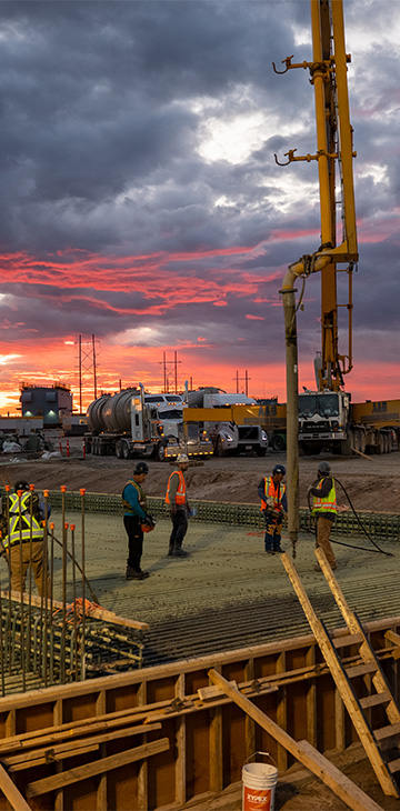 Construction workers on a rebar grid at dusk with a concrete pump and crane under a dramatic sunset sky.